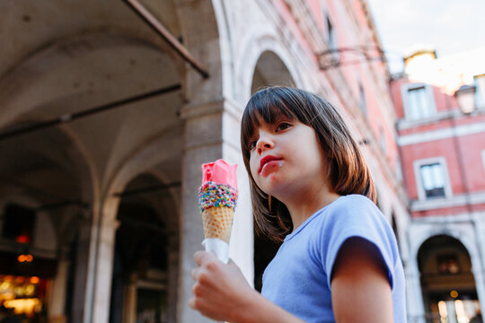Fototapeta Low-angle view of a girl eating ice cream while standing on a street in an Italian town.