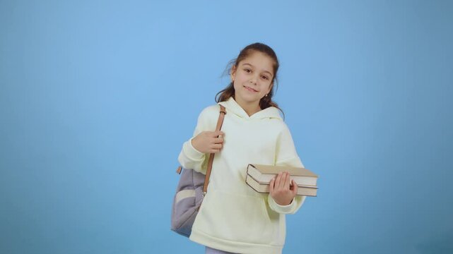 Girl in a light hoodie holds books and gives a thumbs up against a solid blue background in a cheerful indoor setting