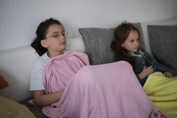 Two sisters snuggled under blankets on the couch watching TV. Concept: sibling bond, cozy evening,...