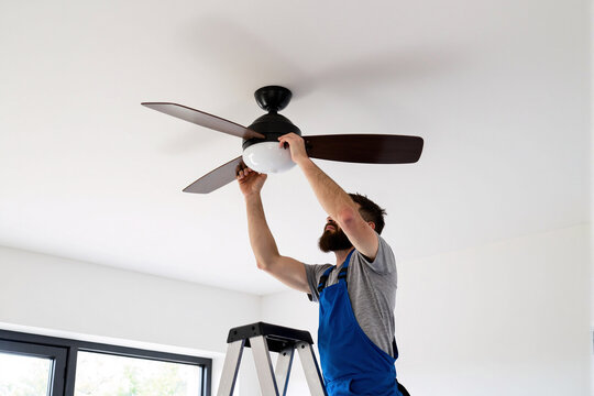Professional electrician in uniform stands on a stepladder to install a modern ceiling fan, performing electrical wiring and home improvement services in a residential room