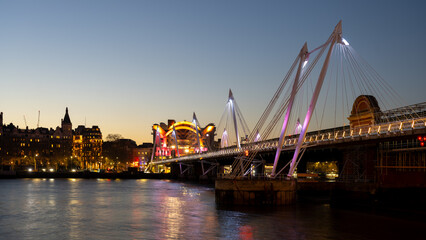The illuminated Charing Cross Bridge spans the River Thames in London during twilight. Surrounding lights reflect on the water, creating a peaceful evening atmosphere for viewers.