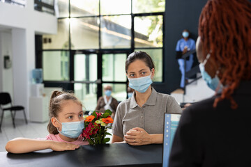 Black receptionist helping mom and child with protective measures in lobby, filling in...