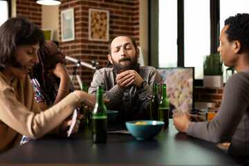 Caucasian man planning his next move during casual poker game with friends at table. Diverse group chatting over cards, beer and snacks, bonding during fun weekend hangout in brick wall room.