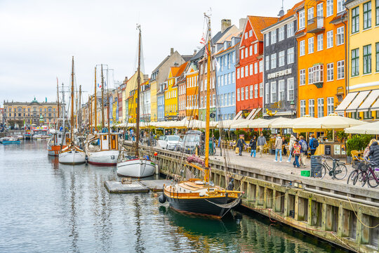 Visitors explore the vibrant Nyhavn area in Copenhagen, Denmark, where historic wooden boats line the canal against a backdrop of colorful buildings and lively cafes.