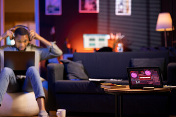 African American man works on laptop at home next to tablet showing alert, notification of a...