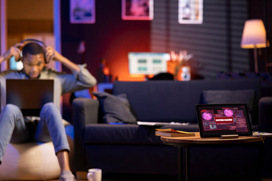 African American man works on laptop at home next to tablet showing alert, notification of a hacking attempt in the cozy living room. Cyberattack and data breach highlight the cyber threat.