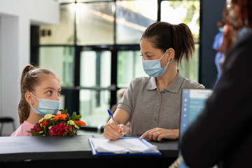 Little girl and mom respecting safety measures with masks at the reception for patient visit, adult...