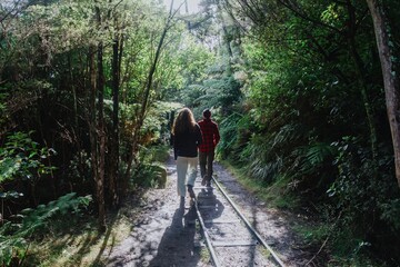 Two people hike along a gravel path with train tracks through a lush forest in The Crown Mines,...
