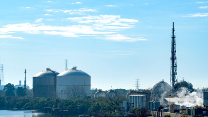 Industrial plant with large storage tanks and chimneys emitting steam, situated near a body of water