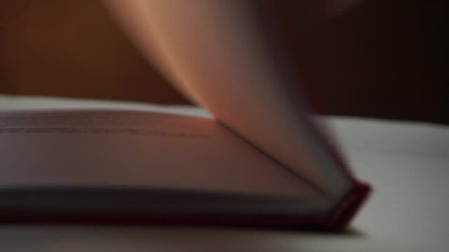 Hand closing an open book with visible red cover, warm light and dark background add mood, suitable for education, ending stories, or thoughtful moments