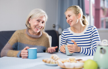 Positive mature woman and her adult daughter, who came to visit her, drink tea and chat, telling...