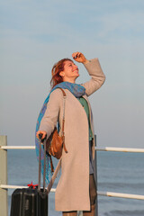 Ginger-haired woman, 30s, standing on pier with suitcase. Happy and stylish in coat and scarf....
