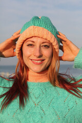 Playful ginger-haired woman, 30s, outdoors by sea, in a colorful outfit and fun hat. Laughing and...