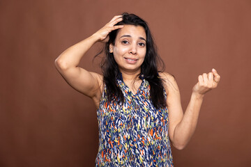 Unsure indian lady stands against brown background, scratching head with puzzled expression. Female...