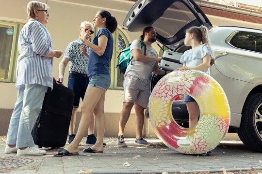 In the driveway, a big family loading their packed car with baggage and folding chair. Grandparents, parents and adorable excited girl prepare for a relaxing summer beach holiday.