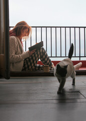 Ginger-haired woman, 30s, sitting on terrace floor in cozy home clothes, reading a book.  Cat...