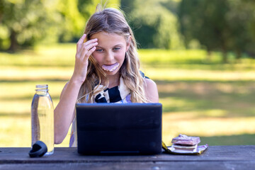 Joyful young girl learning with Dubai chocolate in mouth and laptop on wooden table at sunset. Business laughing woman communicating at homework via wifi on computer with water