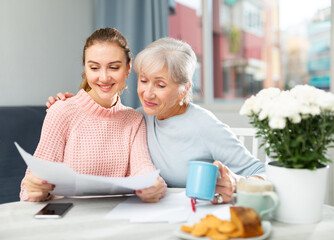 Smiling young girl with elderly mother drinking coffee and reading papers while sitting in cozy...