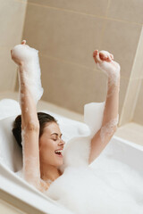 Joyful Caucasian woman enjoying a bubble bath, playfully raising her arms covered in foam....