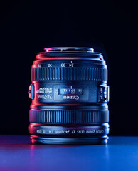 A camera lens stands upright on a dark surface, highlighted by striking blue and pink lights. The...