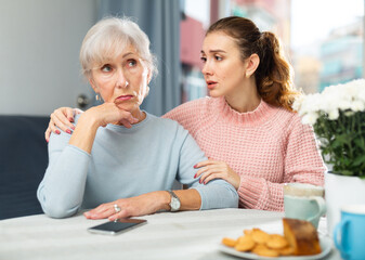 Depressed mature woman sitting at table and her adult daughter trying to encourage her.