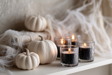 White pumpkins and black candles arranged on a shelf with cobweb decorations. A cozy autumn atmosphere is created with warm candlelight.