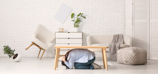 Young woman hiding under table in damaged living room after earthquake