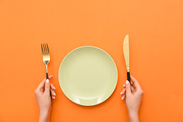 Female hands with cutlery and empty plate on orange background