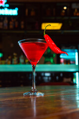 A striking red cocktail with a chili pepper garnish on a wooden bar.