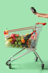 Female hand with payment terminal near shopping cart full of food on green background