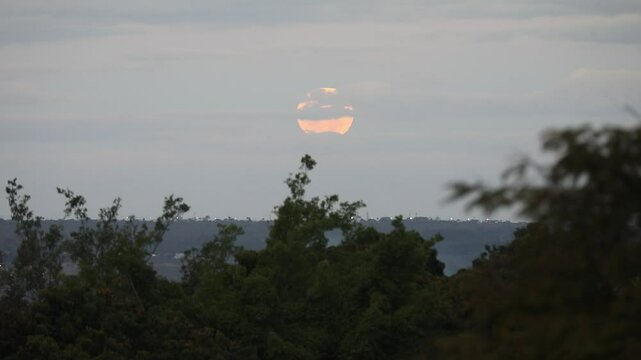 Video of the full moon rising moving through the gray clouds of a dramatic sky, birds flying past in the distance.