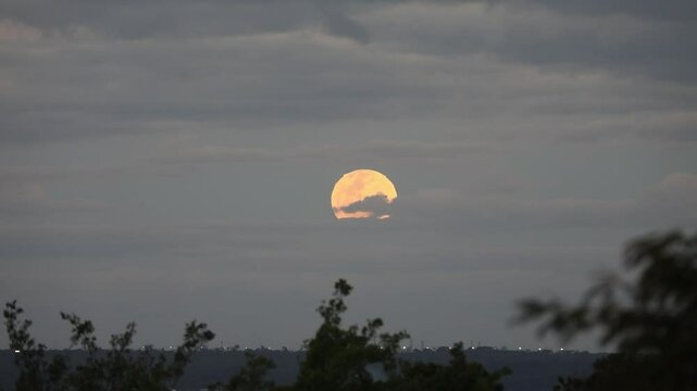 Video of the full moon rising moving through the gray clouds of a dramatic sky, birds flying past in the distance.