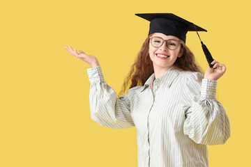Happy female student in graduation hat showing something on yellow background