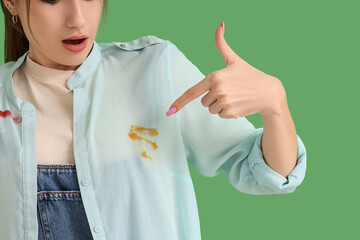 Shocked young woman pointing at stain on dirty shirt against green background