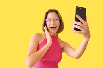 Pretty young woman with mobile phone taking selfie on yellow background