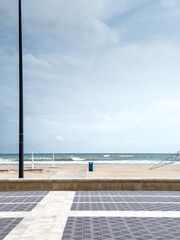 Clouds drift over Platja de la Patacona near Valencia, Spain, where an empty tiled promenade, a lone blue bin and closed showers frame a windswept sandy shore and whitecaps on Mediterranean horizon.