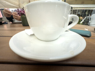 White ceramic coffee cup and saucer photographed in close up on an outdoor wooden cafe table, with blurred patrons, mobile phone and flowers in background, capturing a casual break in daylight.