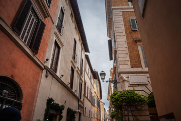 Looking upward along a narrow alley in old Rome where brick facades in italian capital. Tall shuttered windows, wrought iron lamps and creeping greenery evoke the intimate charm of the historic center