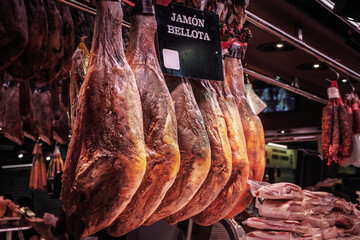 Cured jamon iberico bellota legs hang from metal hooks at a Barcelona market, bright lights highlighting marbled fat and deep red meat of premium Iberico ham, with vacuum-packed slices.