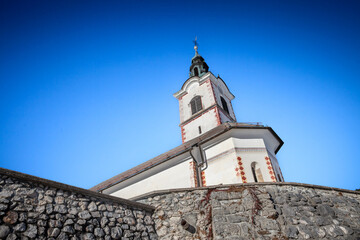 Low angle view of Cerkev Svetega Andreja na Jezerskem shows the white bell tower rising above a stone wall against vivid blue sky, red brick accents and copper spire marking the alpine Catholic landma