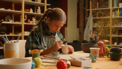 Concentrated talented Caucasian little girl female child schoolgirl in apron daughter kid artist potter holding paint brush painting patterns draw ornament shaping pottery clay pot vase indoor studio
