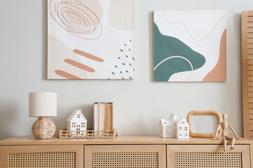 House shaped candle holders, books and lamp on chest of drawers near white wall in room, closeup