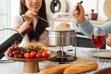 Young happy couple with glass of wine dipping croutons into cheese fondue at home, closeup