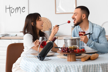 Young happy couple with glasses of wine eating tomatoes dipped in cheese fondue at home