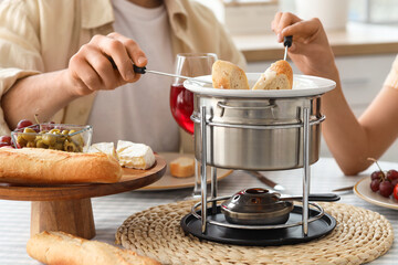 Young couple with glasses of wine dipping croutons into cheese fondue at home, closeup