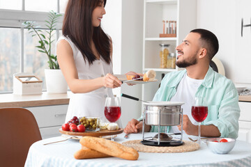 Young happy couple with glasses of wine dipping croutons into cheese fondue at home