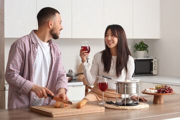 Young happy couple with glasses of wine cutting bread for dipping in cheese fondue at home