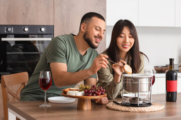 Young happy couple with glasses of wine dipping croutons into cheese fondue at home