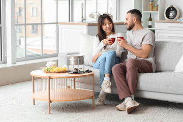 Young happy couple with glasses of wine, sweet snacks and chocolate fondue sitting on sofa at home