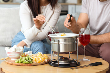 Young couple with glasses of wine dipping sweet snacks into chocolate fondue at home, closeup